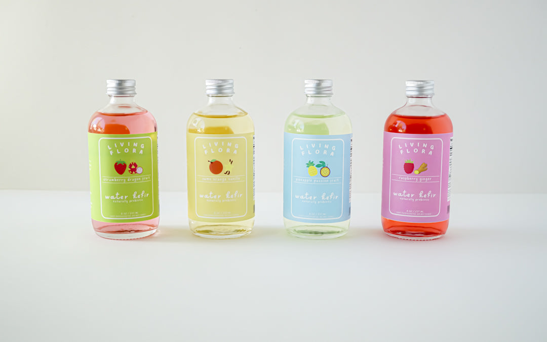 A lineup of 4 water kefir bottles in a row. From left to right, the flavors are strawberry dragon fruit, sumo orange vanilla, pineapple passion fruit, and raspberry ginger.